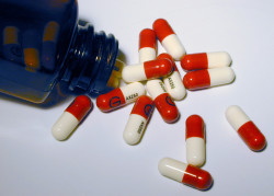 High Angle View of Red and White Medication Capsules Spilling Out of Toppled Glass Bottle on White Background Surface